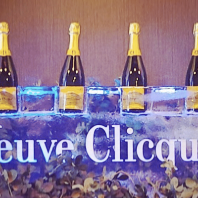 Four bottles of Veuve Clicquot champagne rest in carved compartments atop a clear ice sculpture bearing the Veuve Clicquot name, with decorative greenery at the base.